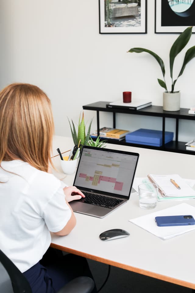 Woman looking at calendar interface on laptop, desk with plants and office supplies in modern workspace.